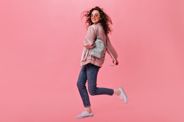 Mujer elegante en jeans y chaqueta moviéndose sobre fondo rosa