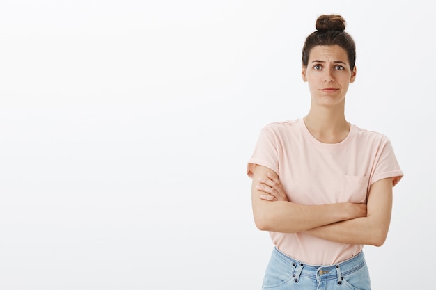 Mujer elegante joven atractiva escéptica que presenta contra la pared blanca