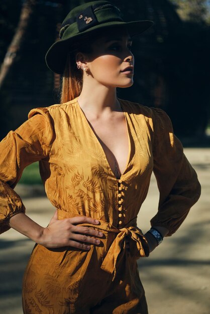 Mujer elegante joven en overoles brillantes con sombrero con confianza mirando hacia otro lado. Hermosa dama posando al aire libre