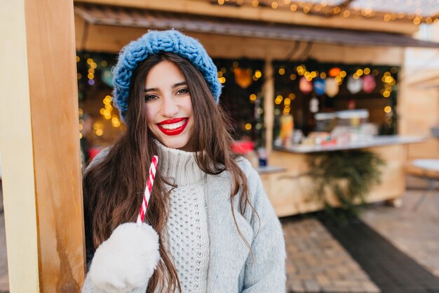 Mujer elegante con maquillaje brillante posando con piruleta cerca del mercado de Navidad en un día frío. La modelo femenina europea complacida viste un abrigo de lana sosteniendo dulces de año nuevo y riendo.