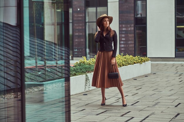 Mujer elegante de moda con chaqueta negra, sombrero marrón y falda con un bolso de mano caminando por el centro de una ciudad europea.