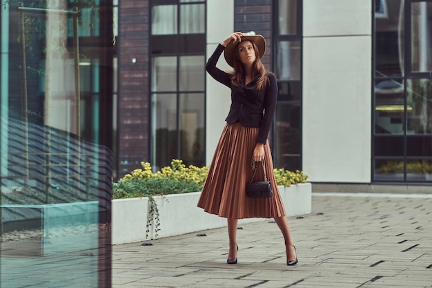 Mujer elegante de moda con chaqueta negra, sombrero marrón y falda con un bolso de mano caminando por el centro de una ciudad europea.