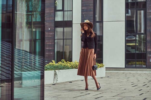 Mujer elegante de moda con chaqueta negra, sombrero marrón y falda con un bolso de mano caminando por el centro de una ciudad europea.