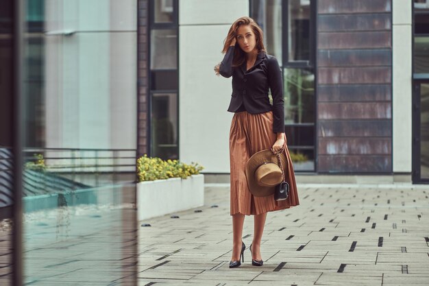 Mujer elegante de moda con chaqueta negra, sombrero marrón y falda con un bolso de mano caminando por el centro de una ciudad europea.