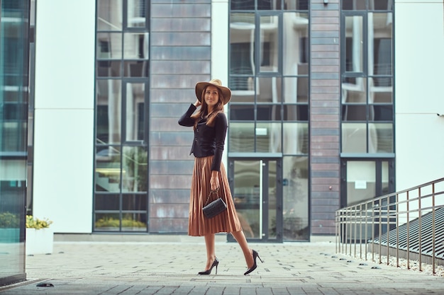 Mujer elegante de moda feliz con una chaqueta negra, un sombrero marrón y una falda con un bolso de mano caminando por el centro de una ciudad europea.