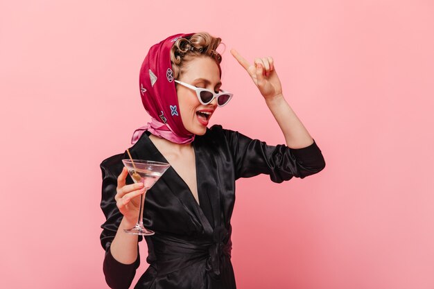 Mujer elegante en pañuelo de seda y vasos con copa de martini en pared rosa