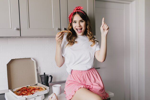 Mujer elegante en pijama romántico comiendo pizza. Encantadora modelo femenina posando en la cocina con una taza de café y comida rápida.