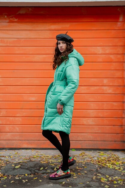 Mujer elegante posando en invierno otoño tendencia de moda abrigo acolchado y sombrero boina contra la pared naranja en la calle vistiendo coloridos zapatos impresos