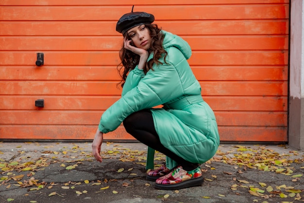 Mujer elegante posando en invierno otoño tendencia de moda abrigo acolchado y sombrero boina contra la pared naranja en la calle vistiendo coloridos zapatos impresos