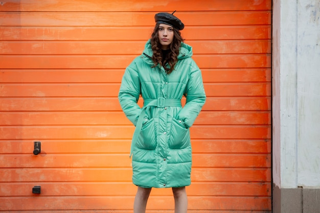 Mujer elegante posando en invierno otoño tendencia de moda abrigo acolchado y sombrero boina contra la pared naranja en la calle