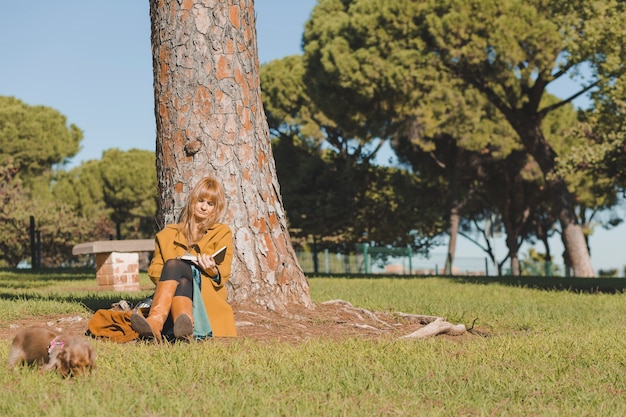 Mujer elegante que descansa con el libro debajo del árbol