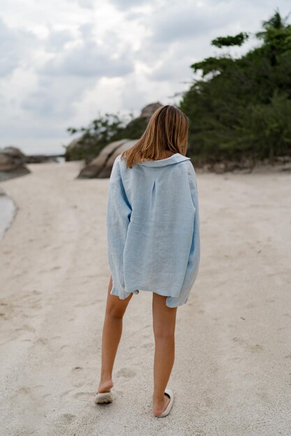 Mujer elegante con ropa casual azul posando en una playa solitaria en una vista nublada de xAweather desde atrás