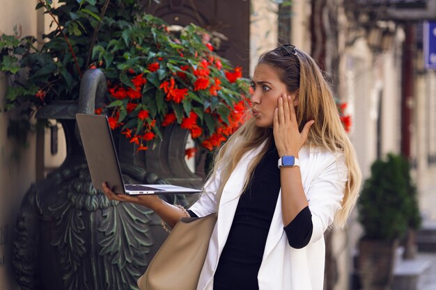 Mujer elegante en ropa de moda mirando en su computadora portátil y se preocupa por algunas noticias. Pone su mano en la mejilla. Reloj en la muñeca, bolso colgando del hombro. De pie junto a flores rojas