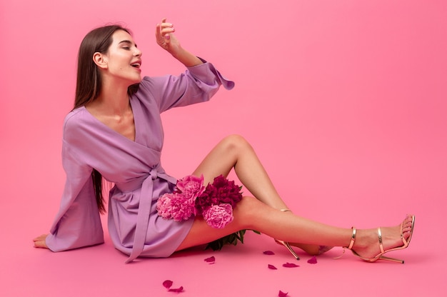 Mujer elegante en rosa en mini vestido de moda de verano violeta posando con ramo de flores de peonía sentado en el piso