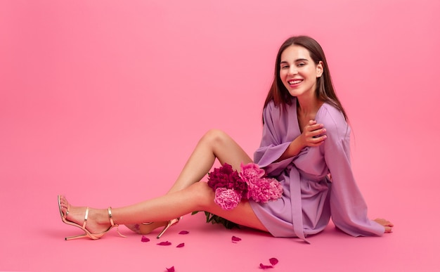 Mujer elegante en rosa en mini vestido de moda de verano violeta posando con ramo de flores de peonía sentado en el piso