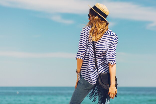 Mujer elegante rubia bronceada bastante delgada con sombrero de paja y gafas de sol, posando en la playa tropical paradisíaca