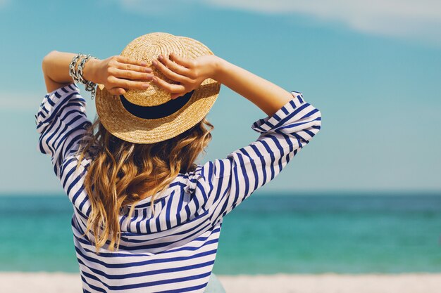 Mujer elegante rubia bronceada bastante delgada con sombrero de paja y gafas de sol, posando en la playa tropical paradisíaca