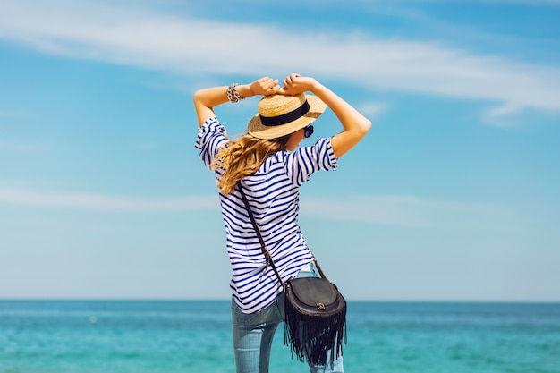 Foto gratuita mujer elegante rubia bronceada bastante delgada con sombrero de paja y gafas de sol, posando en la playa tropical paradisíaca