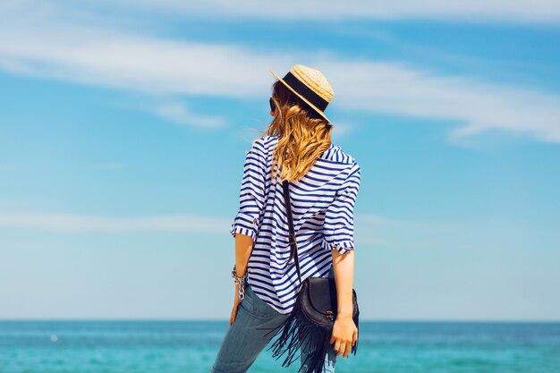 Mujer elegante rubia bronceada bastante delgada con sombrero de paja y gafas de sol, posando en la playa tropical paradisíaca