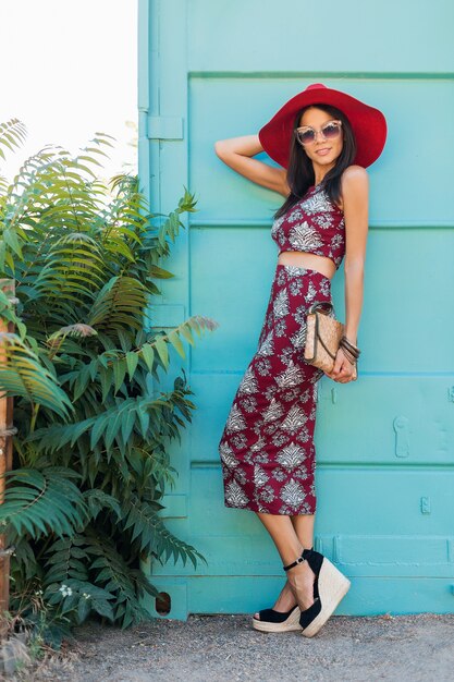 Mujer elegante con sombrero rojo en la pared azul, traje estampado, estilo de verano, tendencia de moda, top, falda, flaco, bolso de paja, gafas de sol, accesorios, sonriendo, vacaciones tropicales, calzado, sandalias en una cuña