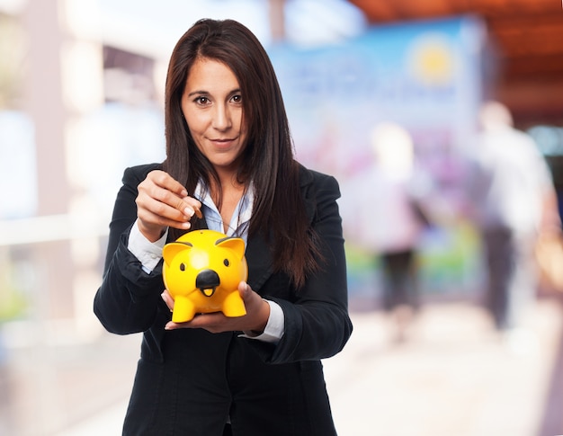 Mujer elegante sonriendo mientras echa una moneda en una hucha amarilla