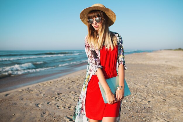 Mujer elegante sosteniendo portátil y caminando en la playa tropical