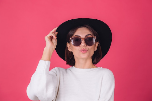 Mujer elegante en suéter blanco casual, gafas de sol y sombrero en la pared rosa roja