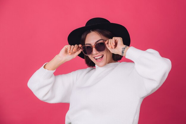 Mujer elegante en suéter blanco casual, gafas de sol y sombrero en la pared rosa roja