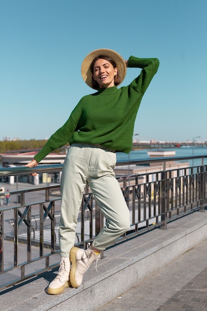 Mujer elegante en suéter casual verde y sombrero al aire libre en el puente con vista al río disfruta de un día soleado de verano
