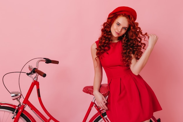 Mujer elegante toca coquetamente su rizo y mira a la cámara, apoyándose en una bicicleta roja en el espacio rosa.