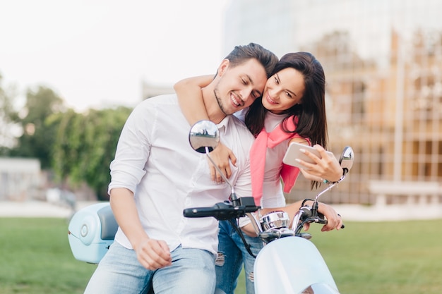 Mujer elegante tocando suavemente a guapo en scooter y haciendo selfie con él