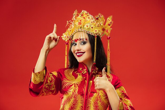 Mujer elegante tradicional china en el estudio sobre fondo rojo. Hermosa chica vestida con traje nacional. Año nuevo chino, elegancia, gracia, ejecutante, actuación, danza, actriz, concepto de vestimenta