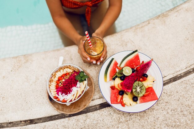 Mujer elegante en traje tropical disfrutando de comida vegetariana. Tazón de batido, plato de fruta y limonada. Vista superior.