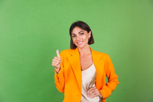 Mujer elegante en vestido beige de seda y blazer naranja de gran tamaño en verde, positivo emocionado muestra el pulgar hacia arriba