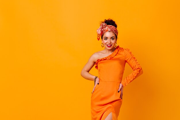 Mujer elegante en vestido de satén naranja y diadema brillante sonriendo y posando en un espacio aislado.