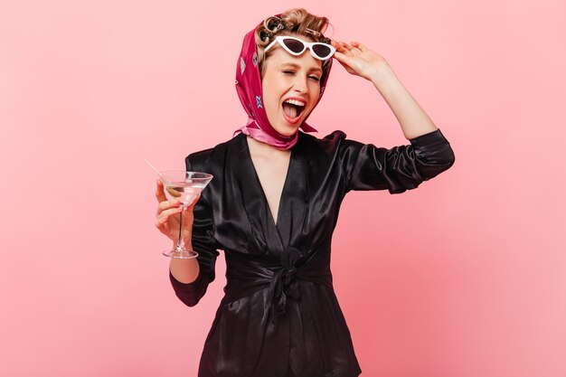 Mujer elegante en vestido de seda y bufanda se quita las gafas, guiña un ojo y posa con martini en la pared rosa