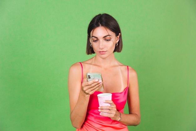 Mujer elegante en vestido de seda rosa aislado en verde feliz con una mirada de sonrisa en la pantalla del teléfono móvil, leer noticias del mensaje