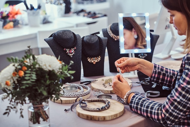 Mujer elegantemente vestida hace collares hechos a mano, trabajando con agujas e hilo en taller de joyería.