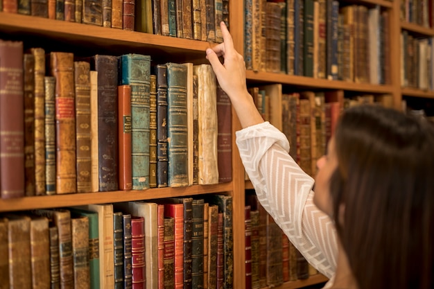 Mujer elegir libro de estantería en biblioteca