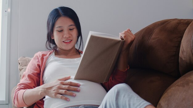 La mujer embarazada asiática joven leyó un libro para el bebé en el vientre. Mamá se siente feliz sonriendo positiva y pacífica mientras cuide al niño acostado en el sofá en la sala de estar en casa.