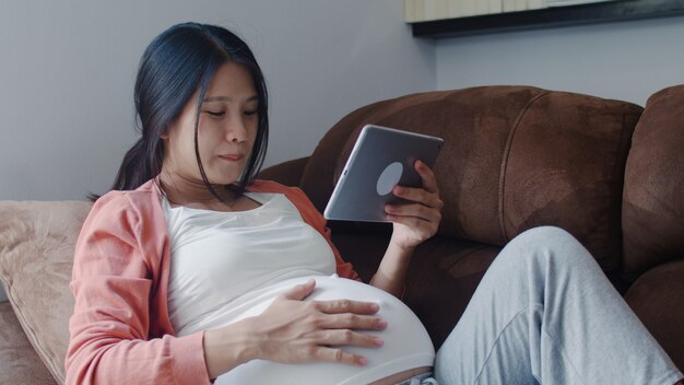 La mujer embarazada asiática joven que usa la tableta busca la información del embarazo. Mamá se siente feliz sonriendo positiva y pacífica mientras cuida a su hijo acostado en el sofá en la sala de estar en casa.