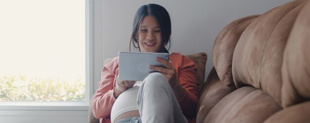 La mujer embarazada asiática joven que usa la tableta busca la información del embarazo. Mamá se siente feliz sonriendo positiva y pacífica mientras cuida a su hijo acostado en el sofá en la sala de estar en casa.