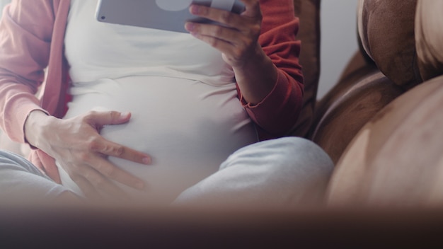 La mujer embarazada asiática joven que usa la tableta busca la información del embarazo. Mamá se siente feliz sonriendo positiva y pacífica mientras cuida a su hijo acostado en el sofá en la sala de estar en casa.