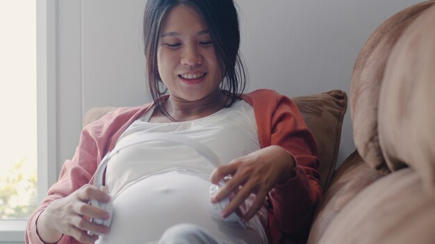 La mujer embarazada asiática joven que usa el teléfono y los auriculares tocan música para el bebé en el vientre. Mamá se siente feliz sonriendo positiva y pacífica mientras cuide al niño acostado en el sofá en la sala de estar en casa.