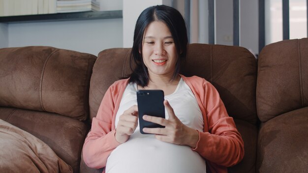 La mujer embarazada asiática joven que usa el teléfono móvil busca la información del embarazo. Mamá se siente feliz sonriendo positiva y pacífica mientras cuida a su hijo acostado en el sofá en la sala de estar en casa.