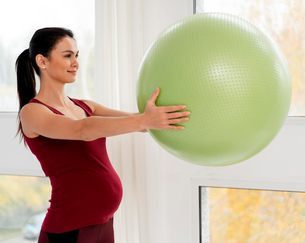 Mujer embarazada de lado sosteniendo una pelota verde fitness