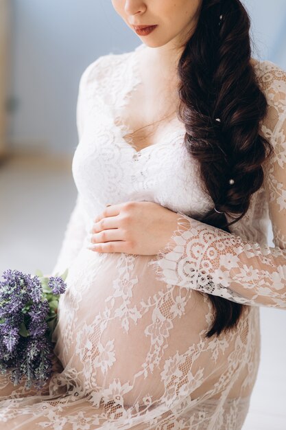 La mujer embarazada magnífica en el vestido blanco presenta en estudio brillante