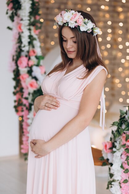 La mujer embarazada morena encantadora en vestido rosado presenta en guirnalda de la flor