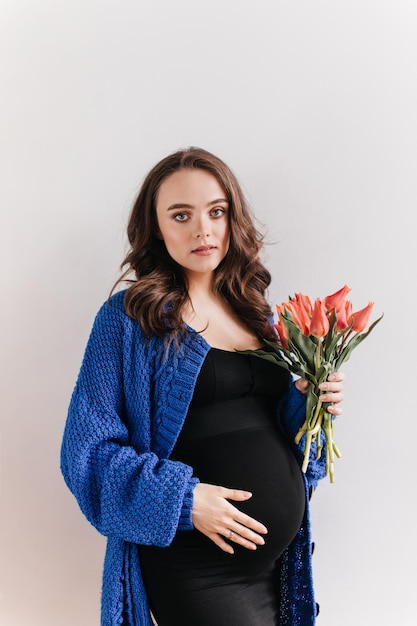 Mujer embarazada morena de ojos azules en vestido negro sostiene tulipanes. Encantadora chica en cardigan posa con ramo de flores sobre fondo blanco.