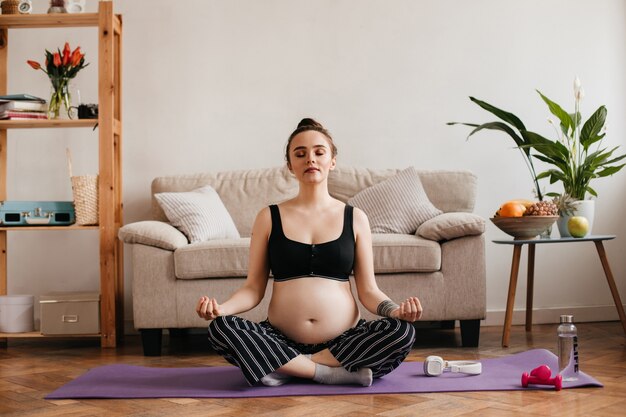 Mujer embarazada en pantalones y top de yoga meditando cerca del sofá beige